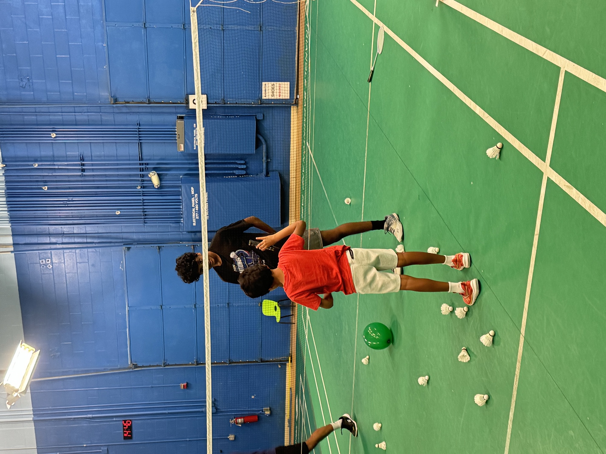 A coach and a young player in a red shirt on a badminton court, with shuttlecocks scattered around them.