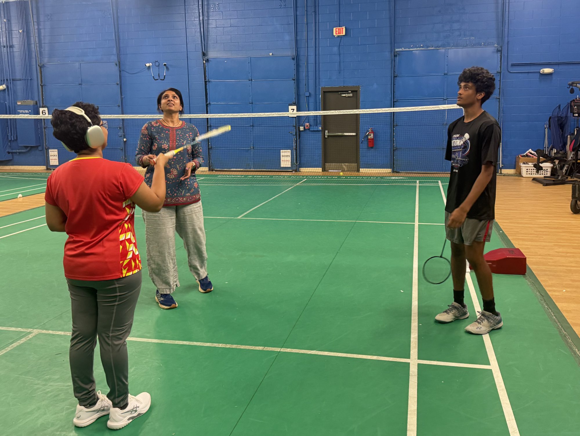 Three players on an indoor badminton court look up at the shuttlecock after a hit.