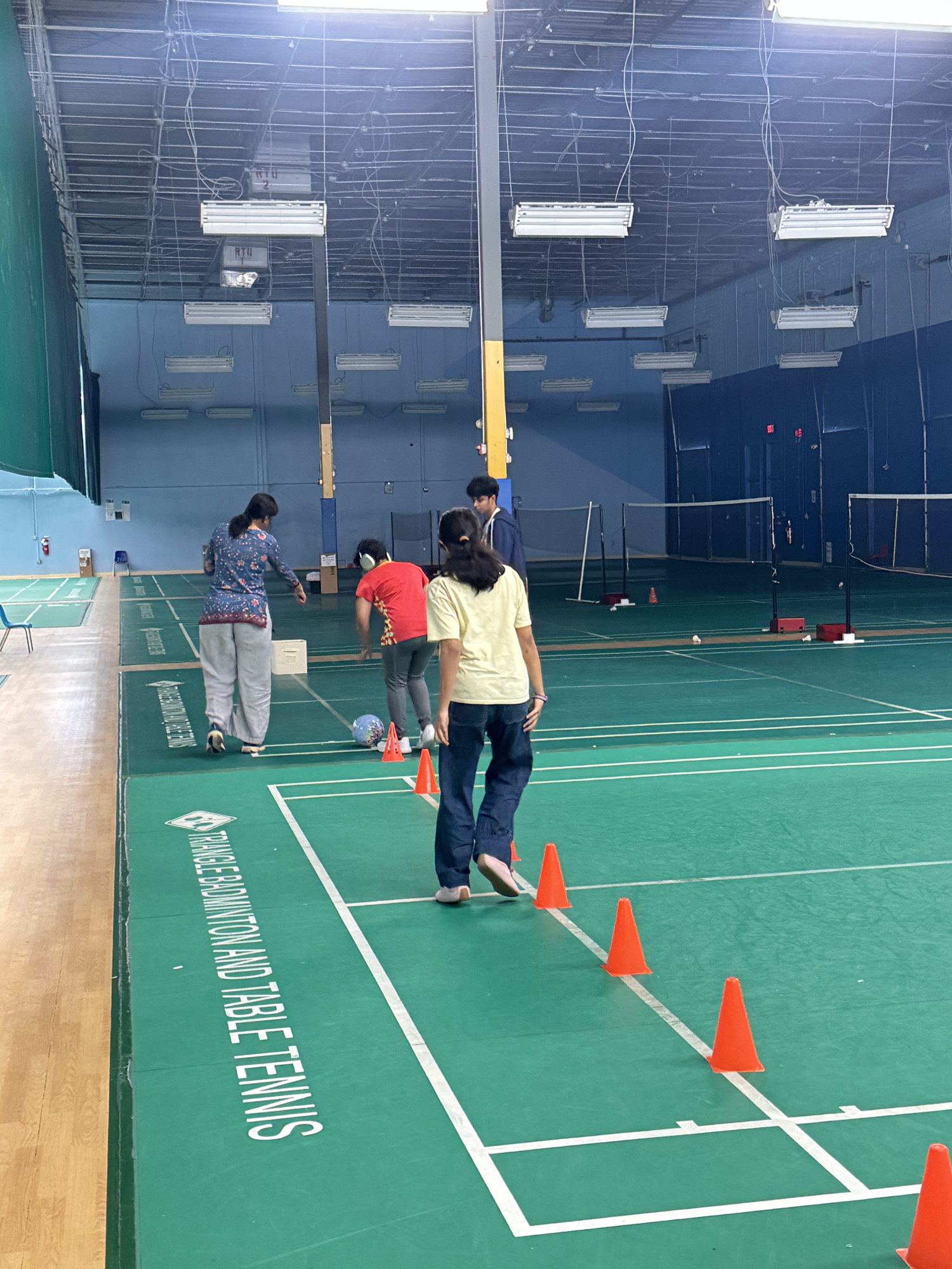 Players weave through a line of orange cones on an indoor badminton court during a warm-up.